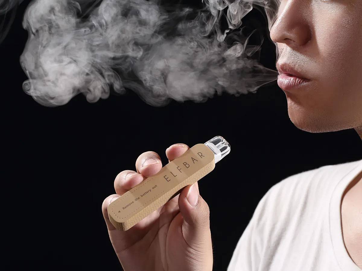 Close-up of a person exhaling vapor while holding a tan ELF BAR disposable vape against a black background.”